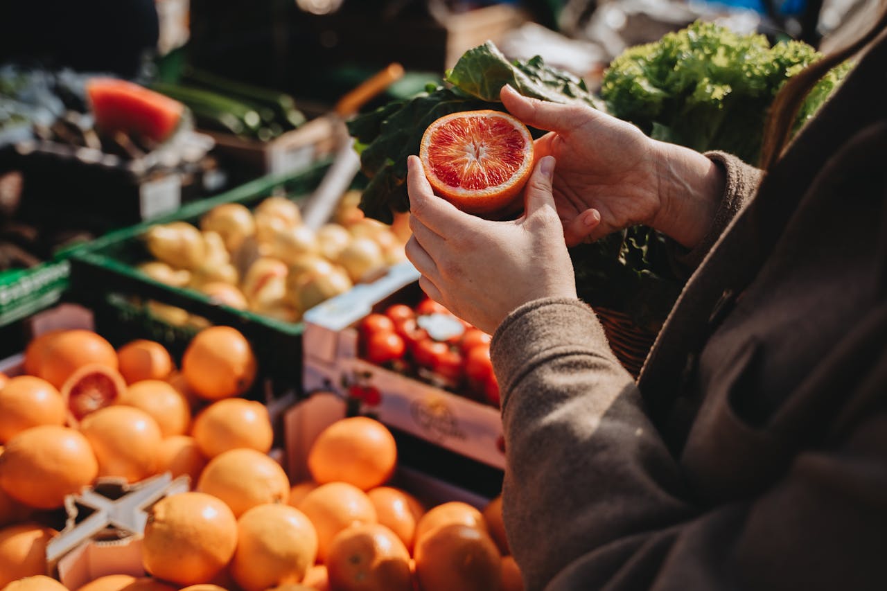 embark A close-up of a person holding a sliced orange at an outdoor market, showcasing fresh produce.